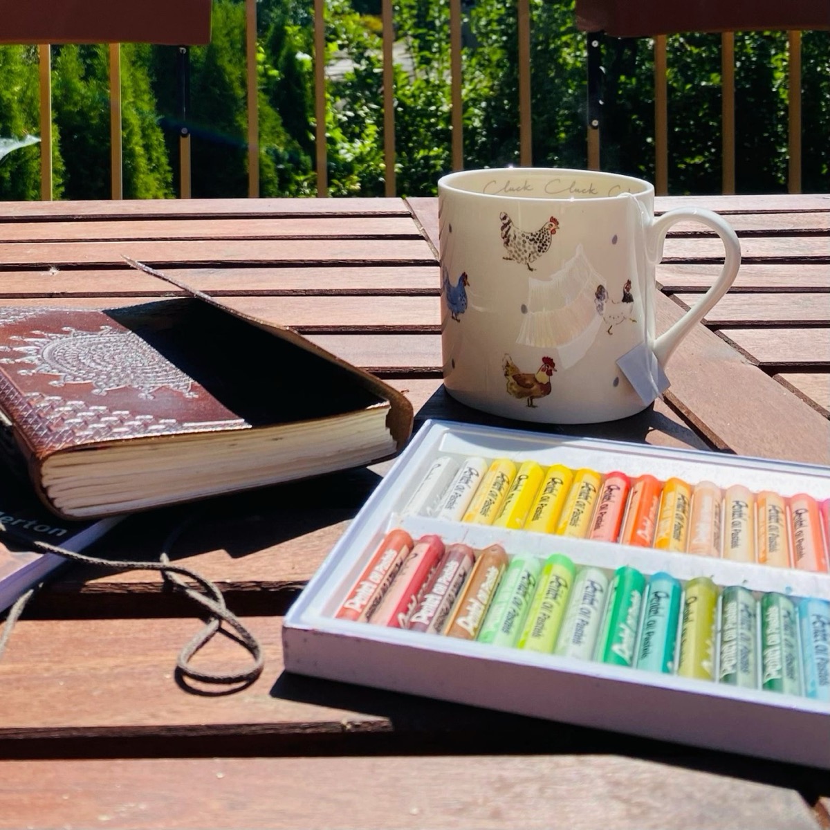 A journal, mug, and box of oil pastels on a sunny outdoor table